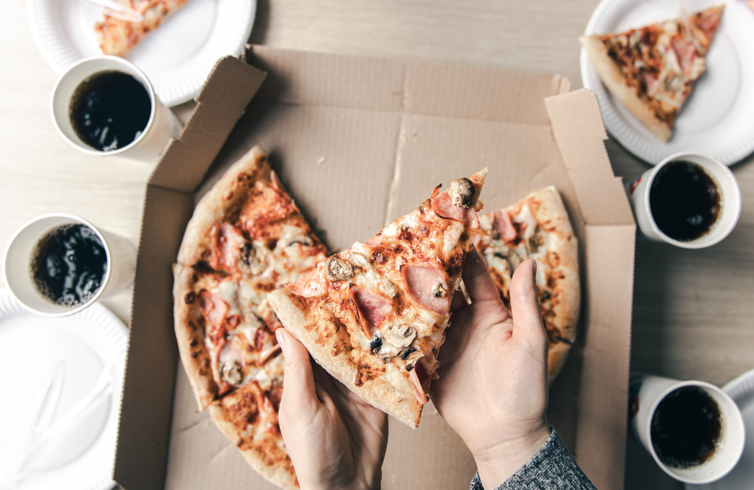 Young woman holding slices of hot pizza from cardboard box at table, top view. Food delivery service.